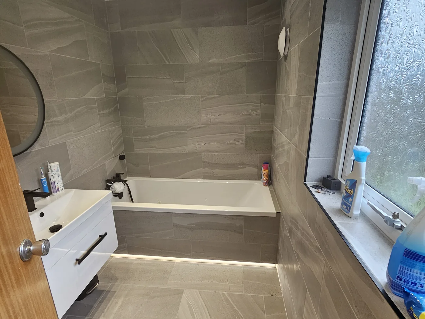 The same bathroom finished in large stone-effect tiles with a bath, vanity unit and lighting under the panel